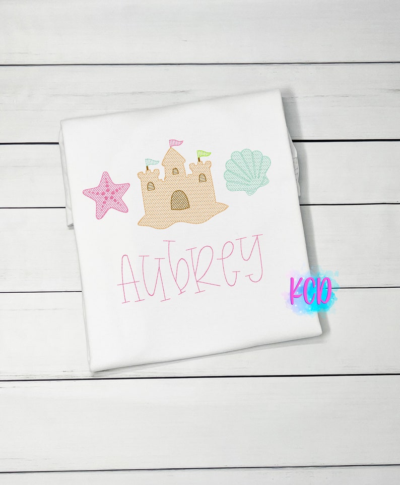 May include: White t-shirt with pink embroidery of a sandcastle, a starfish, and a seashell. The name "Aubrey" is embroidered below the sandcastle.