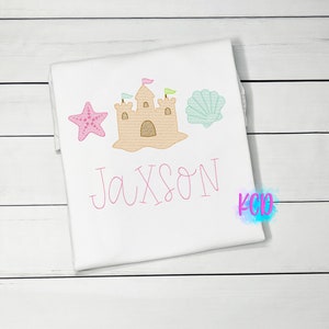 May include: A white t-shirt with a pink sandcastle, a pink starfish, a green seashell, and the name "Jaxson" embroidered in pink.