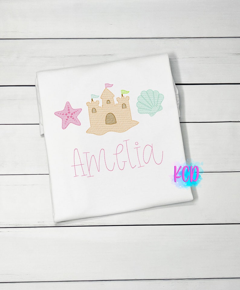 May include: White t-shirt with pink embroidery of a sandcastle, a starfish, and a seashell. The name Amelia is embroidered below the design.