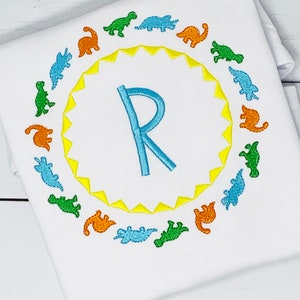 May include: A white onesie with a yellow sunburst circle and a blue letter R in the center.  The circle is surrounded by a ring of colorful dinosaur silhouettes.