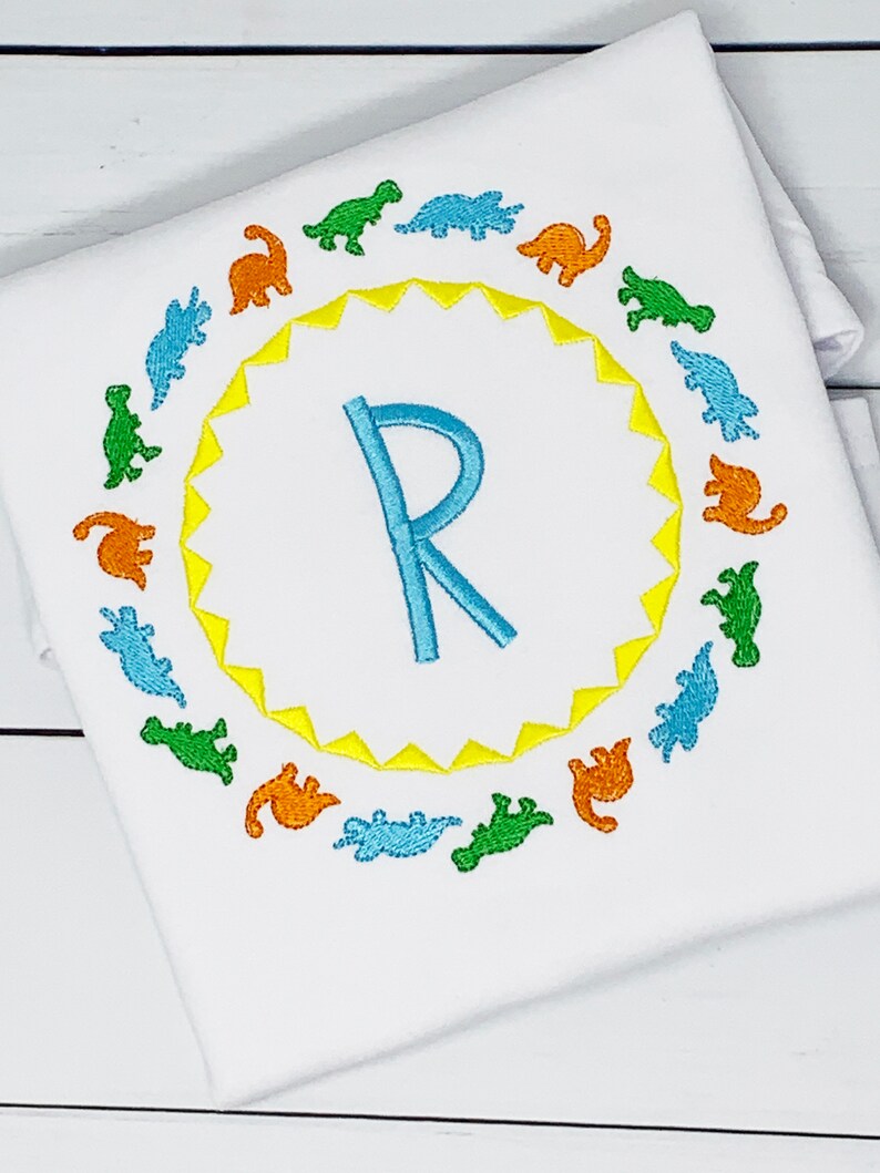 May include: A white onesie with a yellow sunburst circle and a blue letter R in the center.  The circle is surrounded by a ring of colorful dinosaur silhouettes.