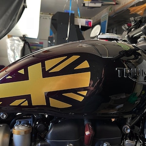 Puede incluir: Un tanque de gasolina de motocicleta negro con una calcoman&iacute;a Union Jack dorada y la palabra "TRIUMPH" en relieve en el lateral.
