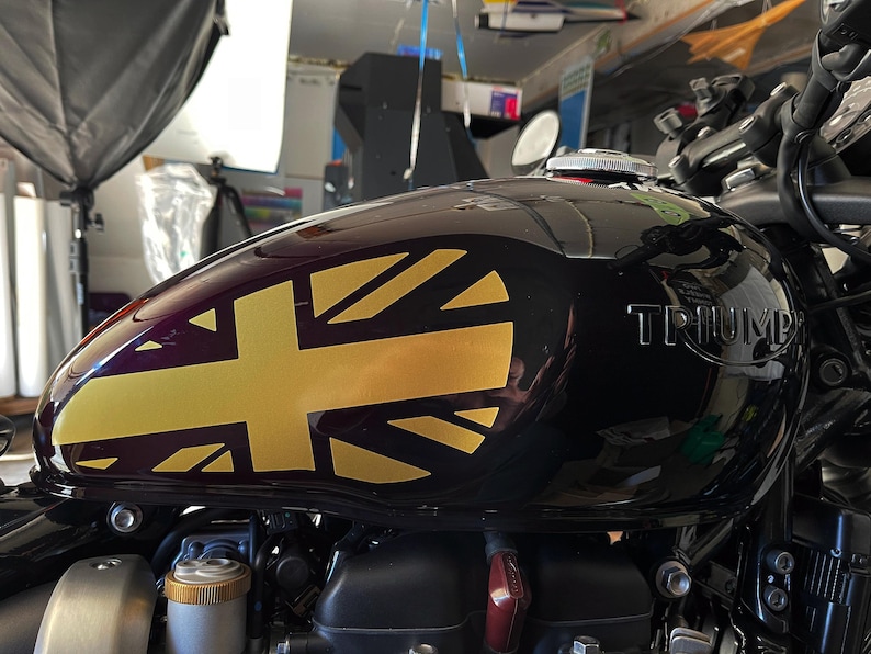Puede incluir: Un tanque de gasolina de motocicleta negro con una calcoman&iacute;a Union Jack dorada y la palabra "TRIUMPH" en relieve en el lateral.