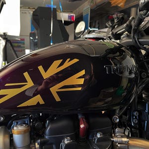 Puede incluir: Un tanque de gasolina de motocicleta negro con una calcoman&iacute;a Union Jack dorada y la palabra "TRIUMPH" en letras plateadas.
