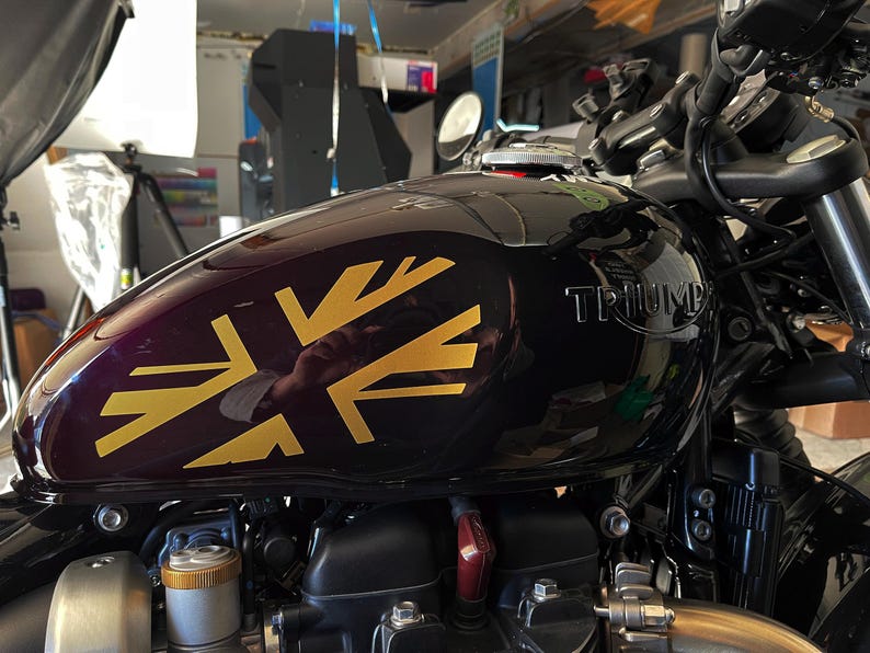 Puede incluir: Un tanque de gasolina de motocicleta negro con una calcoman&iacute;a Union Jack dorada y la palabra "TRIUMPH" en letras plateadas.