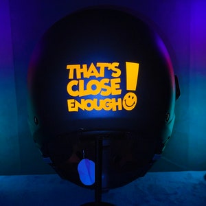 Puede incluir: Casco negro con una pegatina amarilla que dice "That's Close Enough!" y una cara sonriente.