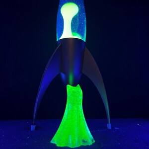 May include: A retro-style lava lamp in the shape of a rocket ship. The lamp features a black base and fins, with a clear glass top containing a green and yellow lava. The lava glows brightly against a dark background.