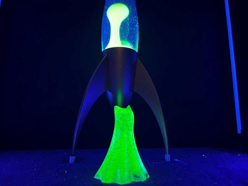 May include: A retro-style lava lamp in the shape of a rocket ship. The lamp features a black base and fins, with a clear glass top containing a green and yellow lava. The lava glows brightly against a dark background.