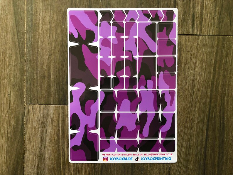 Puede incluir: Una hoja de pegatinas de camuflaje de color morado y negro para personalizar un teclado. Las pegatinas est&aacute;n dispuestas en forma de cuadr&iacute;cula e incluyen teclas de flecha. El texto en la parte inferior dice "WE PRINT CUSTOM STICKERS EMAIL US: HELLO@THEJOYBOX.CO.UK JOYBOXBUDE JOYBOXPRINTING".