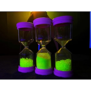 Puede incluir: Tres relojes de arena con tapas y bases moradas, llenos de arena verde brillante. Los cuerpos de vidrio transparente se estrechan en el medio. El fondo es oscuro con un brillo amarillo y azul.
