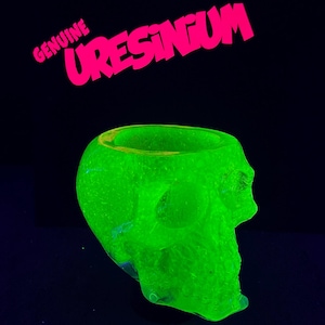 May include: A skull-shaped container glowing in a vibrant neon green color against a black background. The text GENUINE URESINIUM is printed in bright pink letters above the skull. The object features a hollow top and detailed facial features.