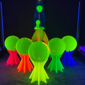 Puede incluir: Una lámpara de lava verde neón con diseño de cohete se encuentra en el centro de la imagen, rodeada de seis orbes verdes brillantes sobre soportes coloridos. Los soportes son rosas, naranjas, verdes y azules, creando una estética retro vibrante.