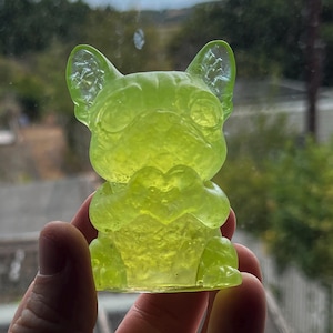 Peut inclure: Une figurine jaune néon translucide en forme de bouledogue français assis. Le chien a de grandes oreilles pointues et tient un petit objet en forme de cœur entre ses pattes avant. Le matériau présente un aspect texturé et cristallin.