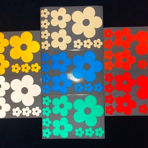 Op de afbeelding: Set van 10 kleurrijke bloemvormige stickers in verschillende kleuren, waaronder geel, wit, blauw, turquoise en rood. De stickers zijn op een grijze achtergrond.