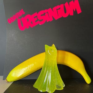May include: A neon green, translucent dress-shaped object stands in front of a yellow banana. The background is black with the words "GENUINE URESINUM" in bright pink.