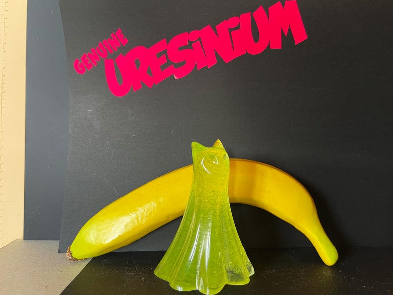 May include: A neon green, translucent dress-shaped object stands in front of a yellow banana. The background is black with the words "GENUINE URESINUM" in bright pink.