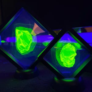 May include: Two black-framed display cases with glowing green objects inside. The objects appear to be carved or shaped, with one resembling a face. The cases are illuminated by blue and green light, creating a vibrant, eye-catching display.