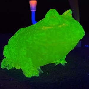 以下が含まれることがあります： ブラックライトで照らされた、鮮やかな緑色のカエルの置物。カエルは座った姿勢で、肌の質感が良く、足と足が見えます。背景は暗く、他のオブジェクトのヒントがあります。
