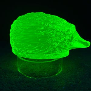 May include: A translucent, bright neon green glass figurine shaped like a hedgehog. The surface features a textured, wavy pattern across the back, and the entire object emits a vivid, glowing light, indicating it is made of uranium glass.