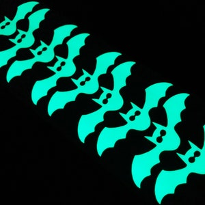 Op de afbeelding: Een rij van acht glow-in-the-dark papieren vleermuizen, elk met een paar ogen, in een verspringend patroon gerangschikt.