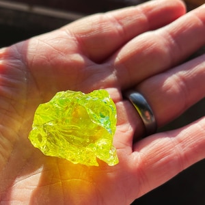Könnte beinhalten: Ein leuchtend gelb-grüner, durchscheinender Glasbrocken mit strukturierter Oberfläche. Die unregelmäßige Form des Glases fängt das Licht ein und erzeugt einen funkelnden Effekt. Das Glas wird in einer offenen Hand gehalten.