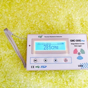 May include: A white GMC-300E Plus Geiger Muller Counter with a digital display showing "281CPM" and the date "01-19-2026". The device has several buttons and is labeled "USA". A lanyard is attached. The background is yellow.