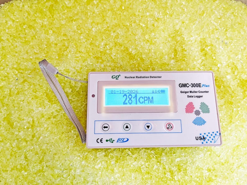 May include: A white GMC-300E Plus Geiger Muller Counter with a digital display showing "281CPM" and the date "01-19-2026". The device has several buttons and is labeled "USA". A lanyard is attached. The background is yellow.