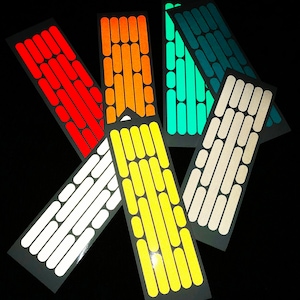 May include: Assortment of six reflective bike spoke stickers in various colors, including white, orange, yellow, green, teal, and beige. Each sticker features a repeating pattern of oval shapes.