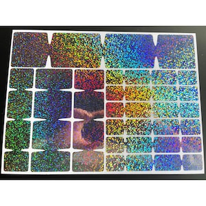 May include: A sheet of iridescent glitter stickers in various shapes and sizes. The stickers have a rainbow effect, with colors shifting from green to blue to gold. The sheet is on a black background.