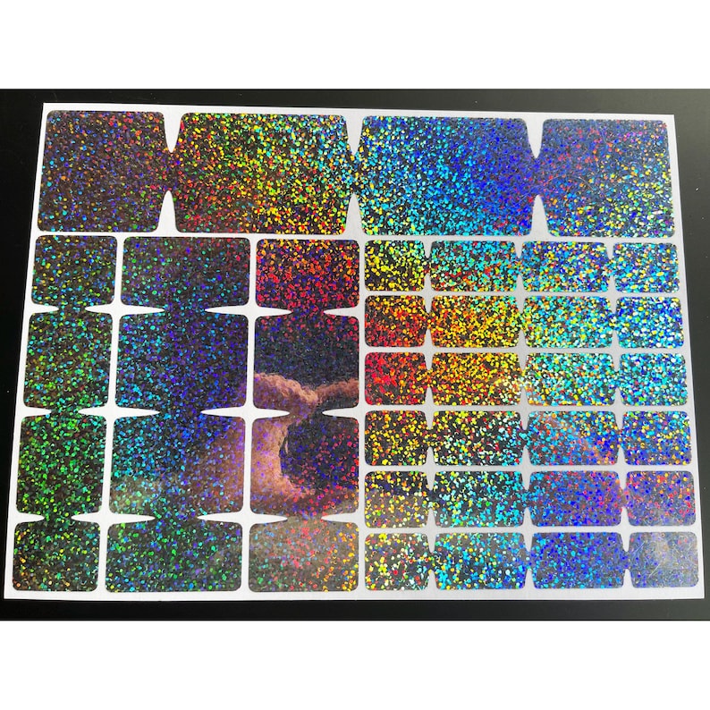 May include: A sheet of iridescent glitter stickers in various shapes and sizes. The stickers have a rainbow effect, with colors shifting from green to blue to gold. The sheet is on a black background.