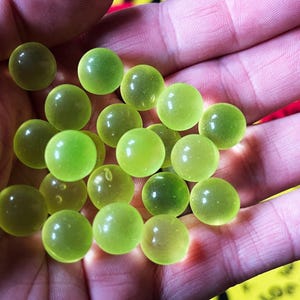 Op de afbeelding: Een handvol doorschijnende, limoengroene knikkers. De bollen variëren enigszins in tint, waarbij sommige geelgroener lijken. De knikkers worden in een hand gehouden, waarbij de huidskleur een contrasterende achtergrond vormt.