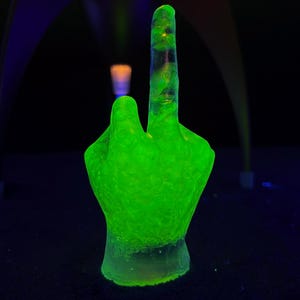 Op de afbeelding: Een neon-groene handsculptuur met de middelvinger omhoog. De sculptuur gloeit helder tegen een donkere achtergrond. De hand is het centrale onderwerp, verlicht met een levendig groen licht.