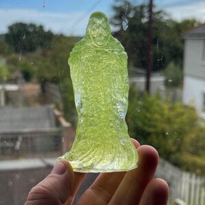 Peut inclure: Une figurine translucide vert citron représentant un personnage squelettique encapuchonné vêtu d'une longue robe fluide. L'objet présente un aspect cristallin texturé et est éclairé par la lumière naturelle, soulignant les détails du crâne.