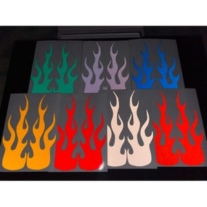 May include: A selection of flame-shaped decals in various colours, including green, purple, blue, yellow, red, and white. The decals are arranged on a dark grey background. These stickers are designed to add a custom look to vehicles or other items.