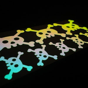 May include: A set of nine iridescent glow-in-the-dark skull and crossbones stickers. The stickers are arranged in a row, with the largest sticker at the top and the smallest sticker at the bottom. The stickers are all different shades of green, yellow, and orange.