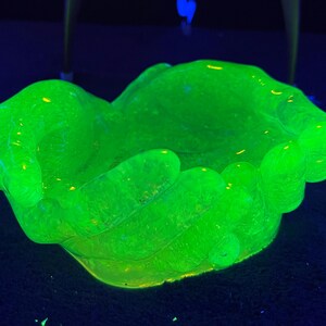 以下が含まれることがあります： 両手を合わせた形をした、輝く緑色のガラスボウル。ボウルは質感のある表面を持ち、暗い青色の背景に対して明るいネオングリーンの光で照らされています。装飾品である可能性が高いです。
