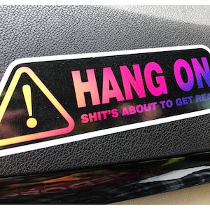 May include: A rectangular sticker with the words "HANG ON" in pink and blue gradient lettering. Below, in smaller text, it reads "SHIT'S ABOUT TO GET REAL." A yellow and black warning symbol is on the left.