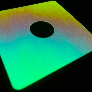 May include: A square, green, yellow, and purple iridescent sticker with a black circle cut out of the center.
