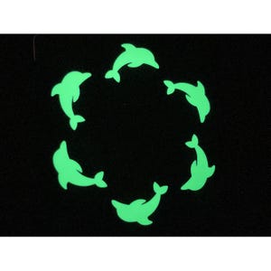 以下が含まれることがあります： 黒い背景に、円形に配置された6つの光る緑色のイルカの形をしたステッカー。ステッカーは均等に配置されており、暗闇で光るように見えます。イルカはそれぞれ異なる方向を向いています。