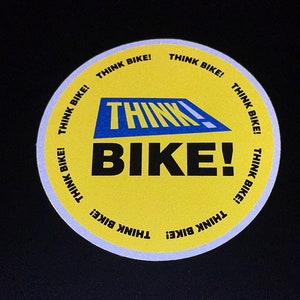 May include: A yellow round sticker with the text "THINK BIKE!" repeated around the edge. In the center, the text "THINK BIKE!" is in black with a blue and white striped rectangle behind it.