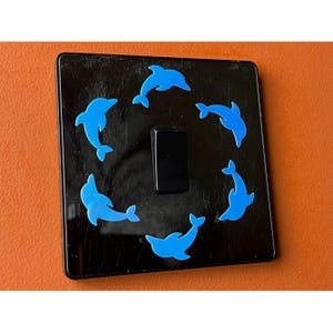 以下が含まれることがあります： 黒いスイッチカバー。6匹の青いイルカのステッカーがスイッチの周りに円を描くように配置されています。イルカは泳いでいるポーズです。スイッチ自体は黒色です。