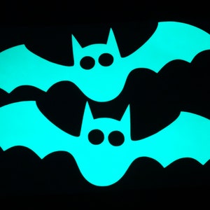 Op de afbeelding: Twee zwarte papieren vleermuizen met een heldere turquoise gloed. De vleermuizen liggen op elkaar, met de bovenste vleermuis iets naar rechts verschoven.