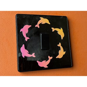 以下が含まれることがあります： ピンクと黄色の色合いの6匹のカラフルなイルカのデカールで飾られた黒いライトスイッチカバー。イルカはスイッチの周りに円を描くように配置されています。カバーは正方形で、光沢のある仕上がりです。
