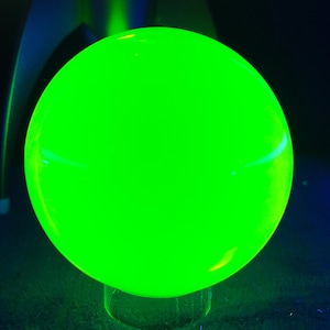Könnte beinhalten: Eine leuchtende, leuchtend grüne Kugel steht im Mittelpunkt. Die Kugel, möglicherweise aus Glas oder Kunststoff, ist hell beleuchtet und erzeugt ein starkes Leuchten. Der Hintergrund ist dunkel, mit Hauch von Blau und Grün.