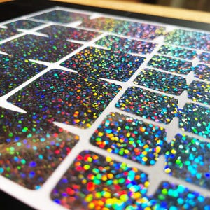 May include: Close-up of a sheet of iridescent glitter stickers. The stickers are rectangular with rounded corners and are arranged in a grid pattern. The glitter reflects a rainbow of colors, including blue, green, yellow, and red.
