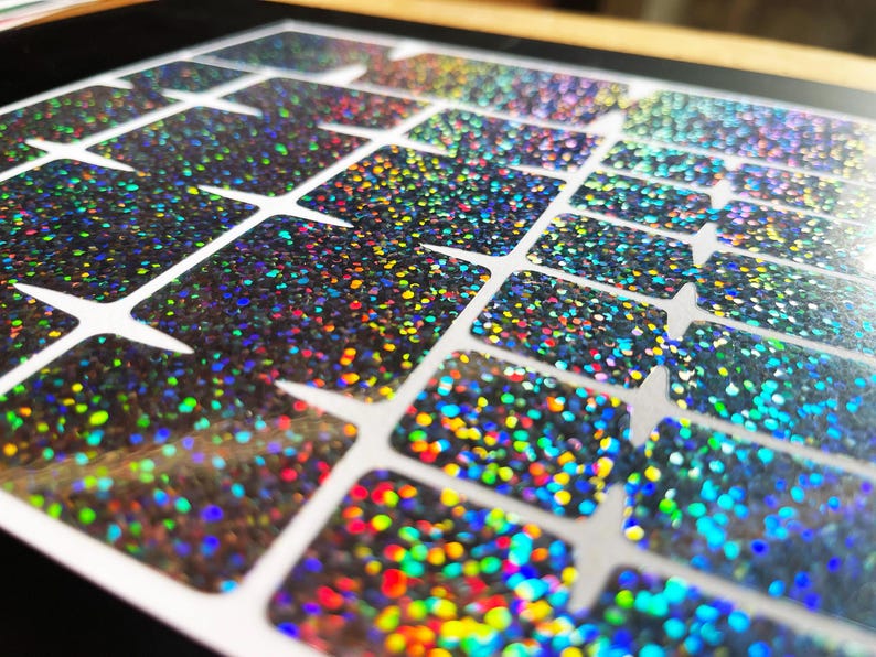 May include: Close-up of a sheet of iridescent glitter stickers. The stickers are rectangular with rounded corners and are arranged in a grid pattern. The glitter reflects a rainbow of colors, including blue, green, yellow, and red.