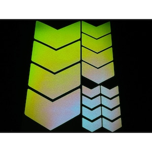 May include: A collection of reflective chevron-shaped stickers in various sizes. The stickers transition in color from green to silver, with a black background. These stickers are designed for enhanced visibility.