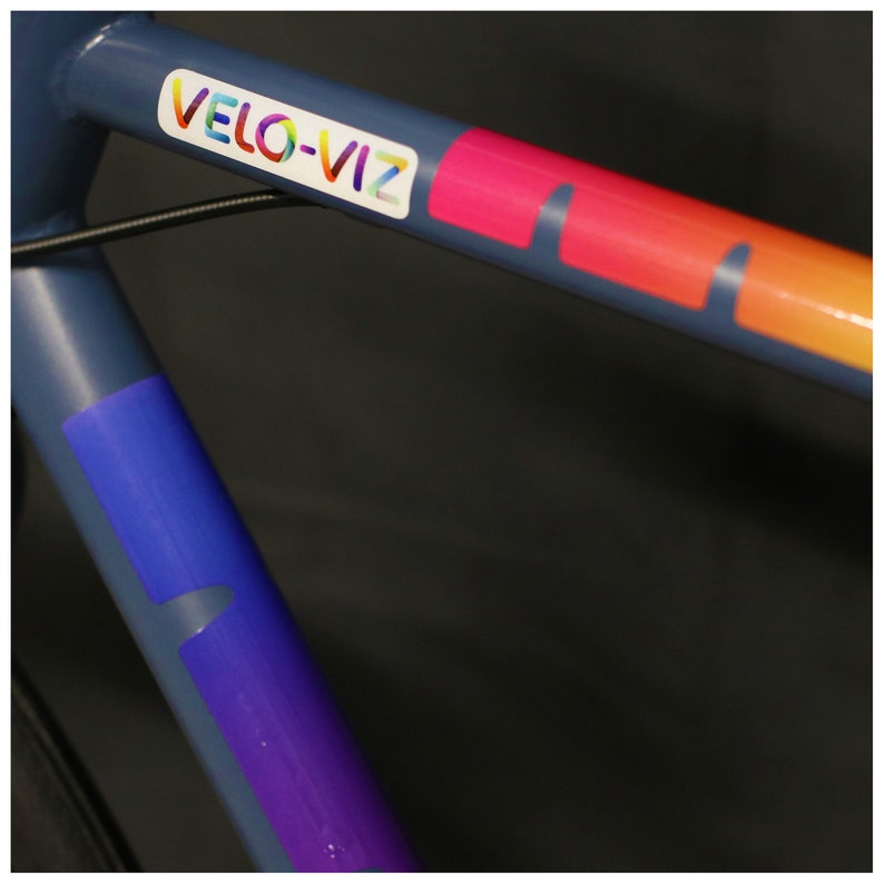 May include: A close-up of a blue bicycle frame with a colorful "VELO-VIZ" sticker and a purple and blue stripe.