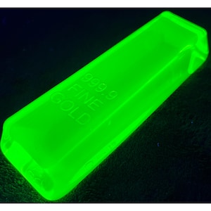 May include: A luminous green rectangular bar with rounded edges. The text "999.9 FINE GOLD" is embossed on the surface. The bar is illuminated against a dark background, creating a striking contrast.