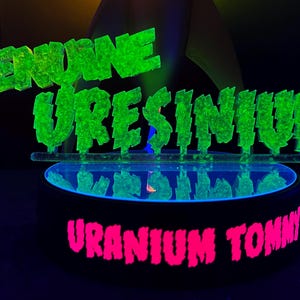 May include: A neon green sign that reads "GENUINE URESINIUM" above a black circular base with the words "URANIUM TOMMY" in pink. The sign and base are illuminated with a black light, creating a glowing effect.